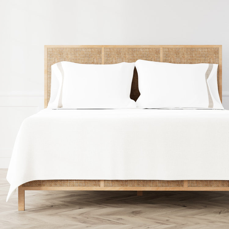A neatly made bed featuring the Minaudiere Sheet Set in White‑Taupe from the Judith Leiber Couture by Ann Gish collection, styled against a light wood rattan headboard and matching footboard in a bright, minimalist room. - SSMIK-WHI-TAU SSMIQ-WHI-TAU