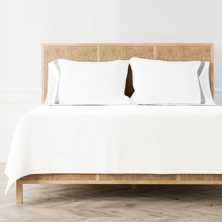 A neatly made bed featuring the Minaudiere Sheet Set in White‑Metal from the Judith Leiber Couture by Ann Gish collection, styled against a light wood rattan headboard and matching footboard in a bright, minimalist room. - SSMIK-WHI-MET SSMIQ-WHI-MET