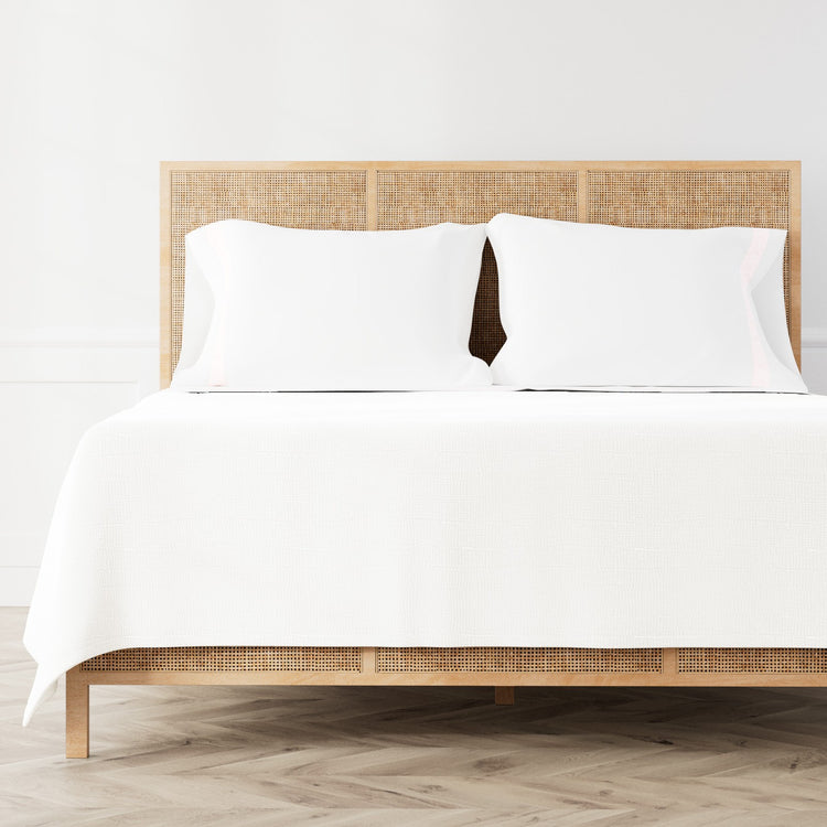 A neatly made bed featuring the Minaudiere Sheet Set in White‑Pink from the Judith Leiber Couture by Ann Gish collection, styled against a light wood rattan headboard and matching footboard in a bright, minimalist room. - SSMIK-WHI-PIN SSMIQ-WHI-PIN