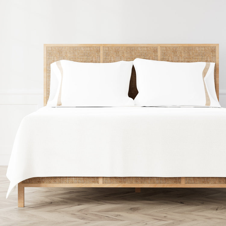 A neatly made bed featuring the Minaudiere Sheet Set in White‑Sand from the Judith Leiber Couture by Ann Gish collection, styled against a light wood rattan headboard and matching footboard in a bright, minimalist room. - SSMIK-WHI-SAN SSMIQ-WHI-SAN