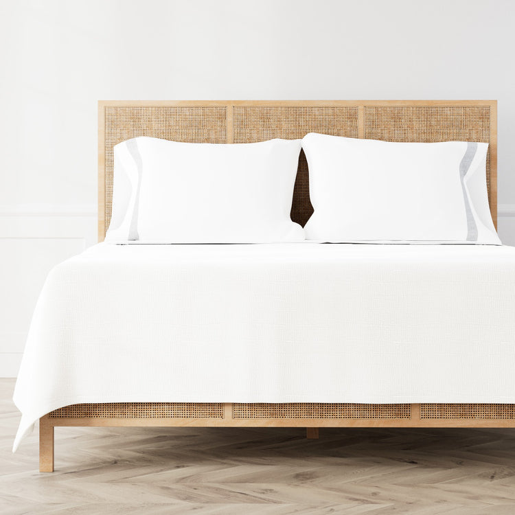 A neatly made bed featuring the Minaudiere Sheet Set in White‑Silver from the Judith Leiber Couture by Ann Gish collection, styled against a light wood rattan headboard and matching footboard in a bright, minimalist room. - SSMIK-WHI-SIL SSMIQ-WHI-SIL