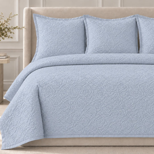 The Roses Coverlet Set from Judith Leiber Couture by Ann Gish, shown in Blue, featuring its signature rose‑quilted pattern with matching shams, styled on a bed with a beige upholstered headboard in a softly lit, paneled bedroom. - YSETCORQK‑BLE YSETCORQQ‑BLE 