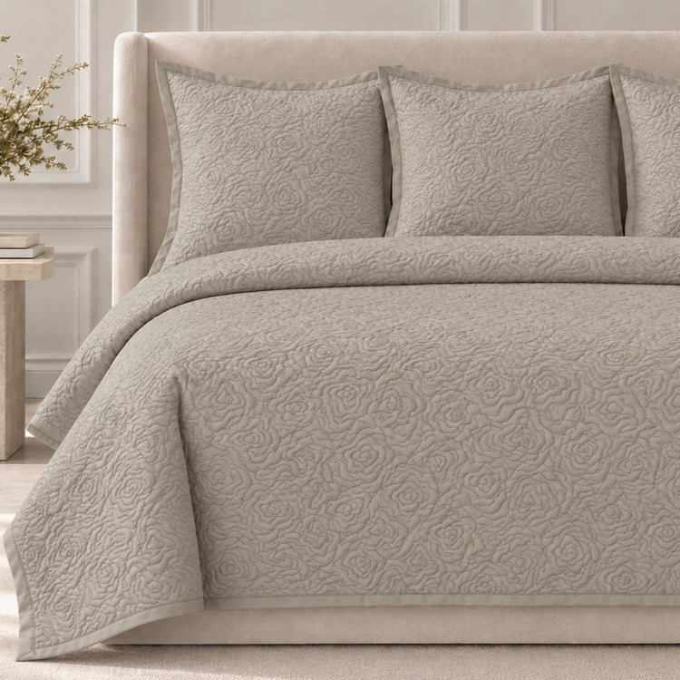 The Roses Coverlet Set from Judith Leiber Couture by Ann Gish, shown in Taupe, featuring its signature rose‑quilted pattern with matching shams, styled on a bed with a beige upholstered headboard in a softly lit, paneled bedroom. - YSETCORQK‑TAU YSETCORQQ‑TAU