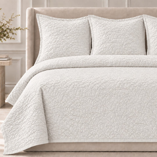 The Shells Coverlet Set from the Judith Leiber Couture by Ann Gish collection, shown in white with its scalloped quilted pattern, styled on a bed with a beige upholstered headboard and matching shams in a softly lit bedroom. - YSETCOSHK-WHI YSETCOSHQ-WHI